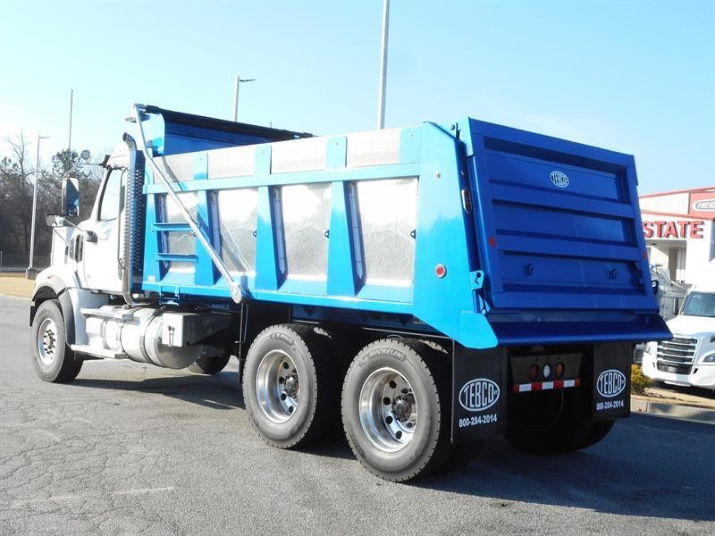 Dump truck rear and side view showing Western Star 47X dump body, blue cargo bed, dual rear wheels, white cab, visible tires and registration stickers.