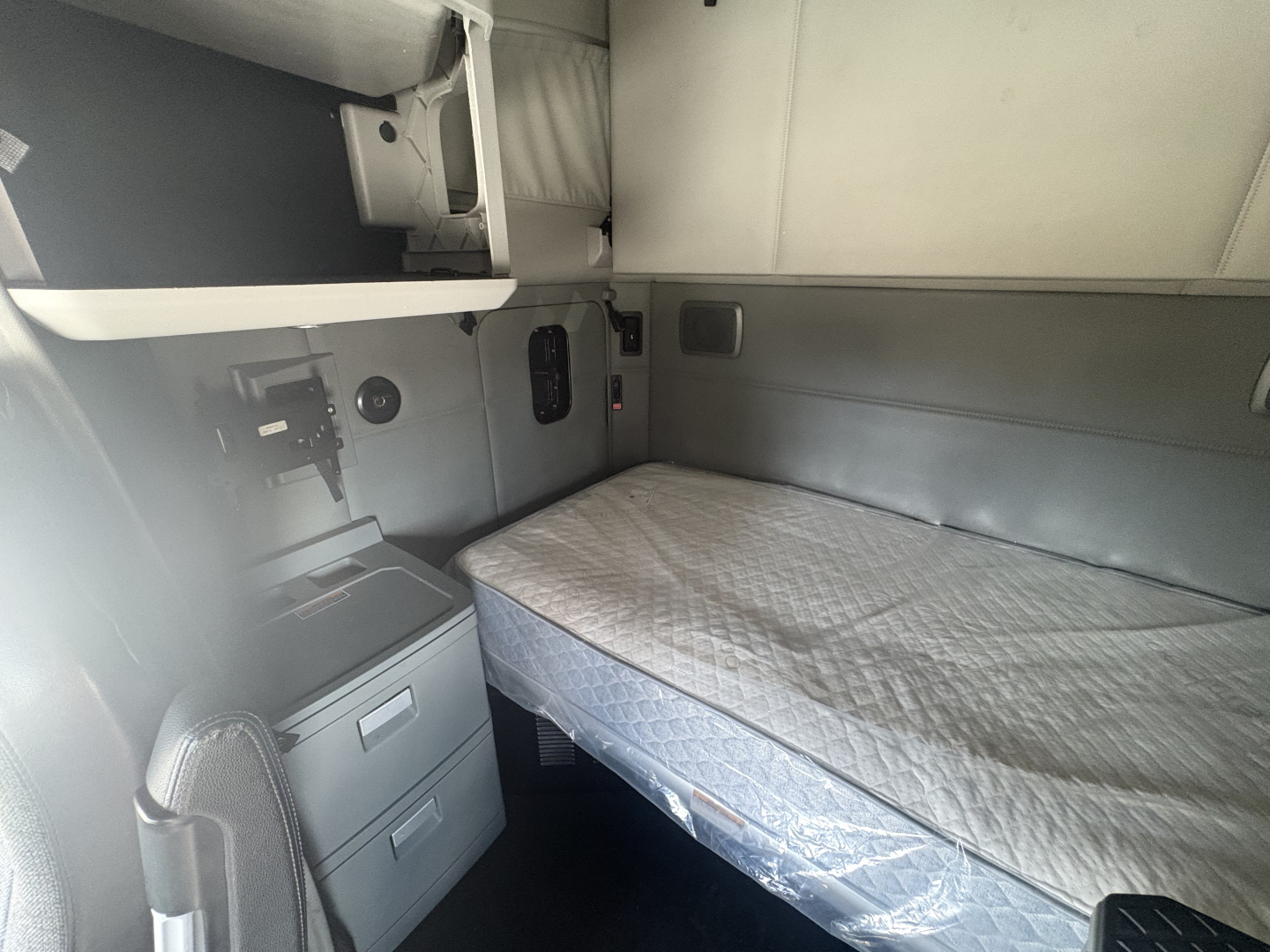 Kenworth T680 sleeper cab interior showing bed with mattress, overhead cabinets, and control panel area on left side.