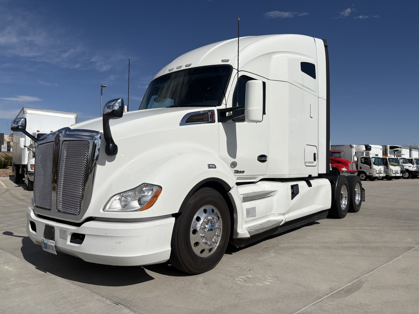 Kenworth T680 tractor unit in white with sleeper cab, front left view, chrome grille, multiple axles, trucks in background.