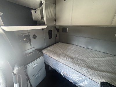 Kenworth T680 sleeper cab interior showing bed with mattress, overhead cabinets, and control panel area on left side.