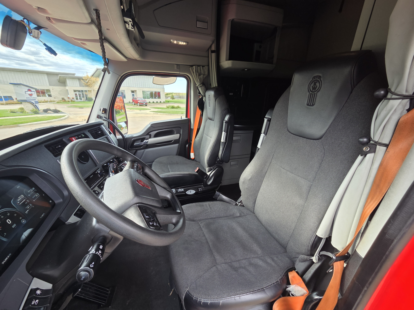Kenworth T680 cabin interior showing steering wheel, driver's seat, and dashboard with gray fabric seats and overhead bunk area.