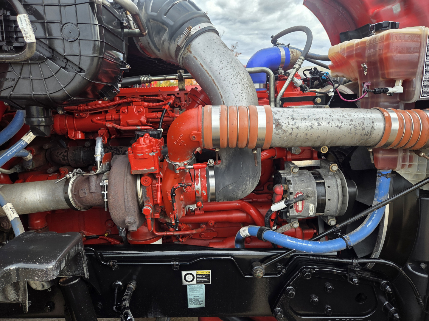 Kenworth T680 engine bay showing red engine components, large silver intake pipe with orange couplings, blue hoses, and coolant reservoir.