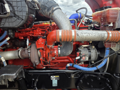 Kenworth T680 engine bay showing red engine components, large silver intake pipe with orange couplings, blue hoses, and coolant reservoir.