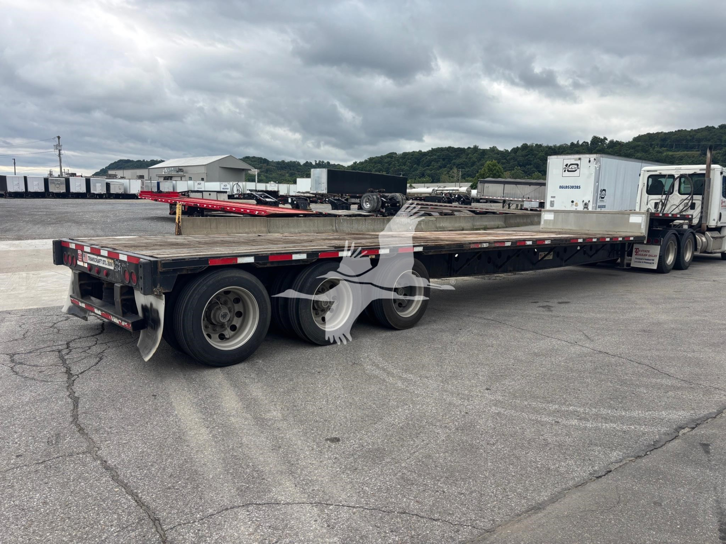 Flatbed trailer (2015 Transcraft DTL-3000) showing long low-bed with dual rear axles and a white cab truck in background.