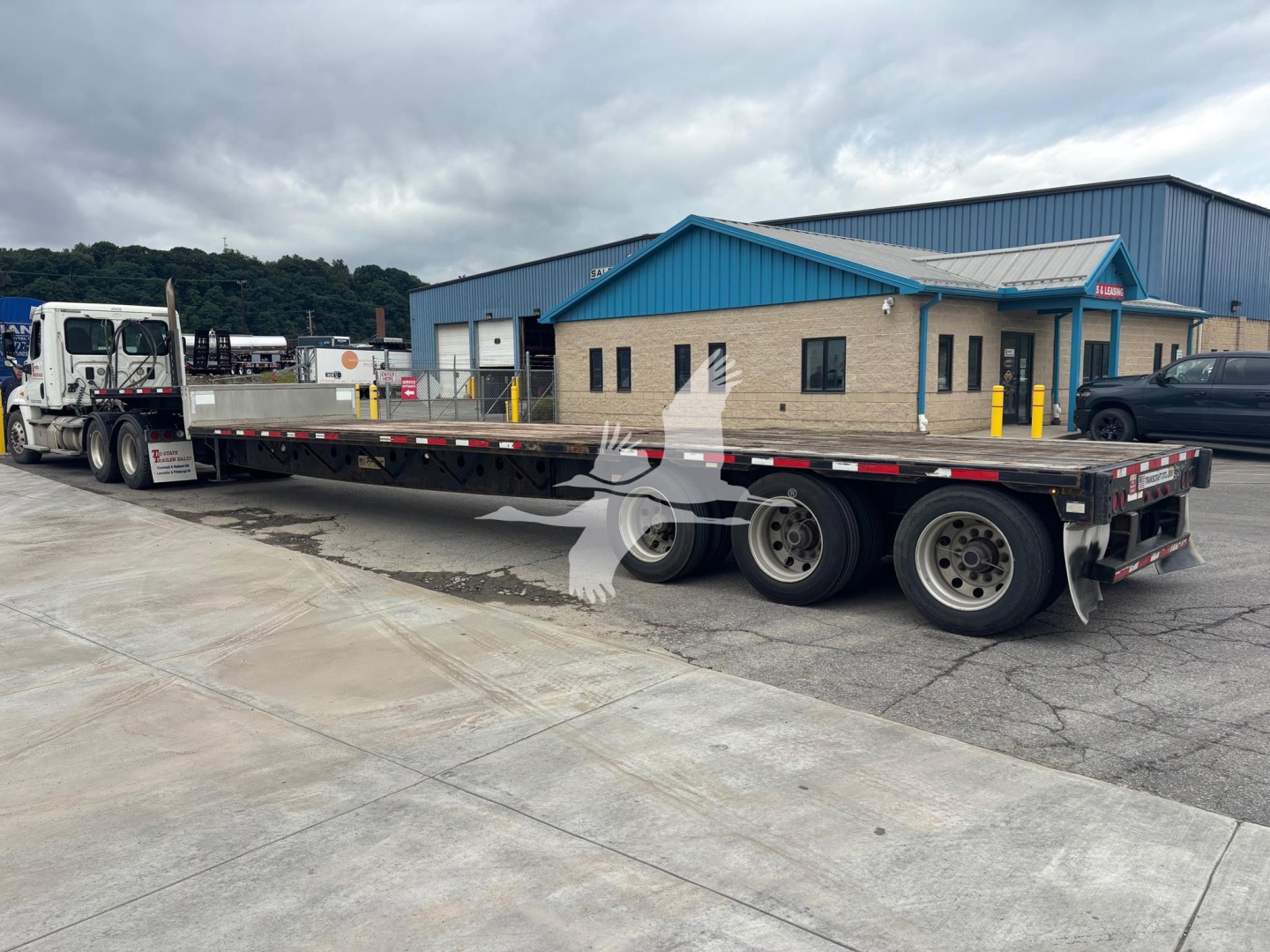 Transcraft DTL-3000 drop deck trailer showing long flatbed with three axles and dual rear braking system, with a blue-roof building in background.