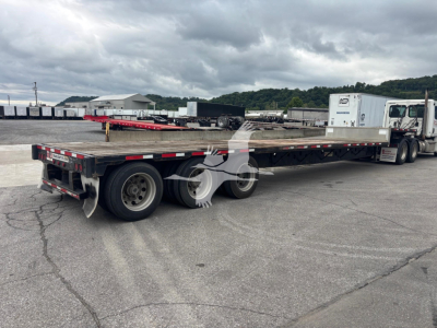 Flatbed trailer (2015 Transcraft DTL-3000) showing long low-bed with dual rear axles and a white cab truck in background.