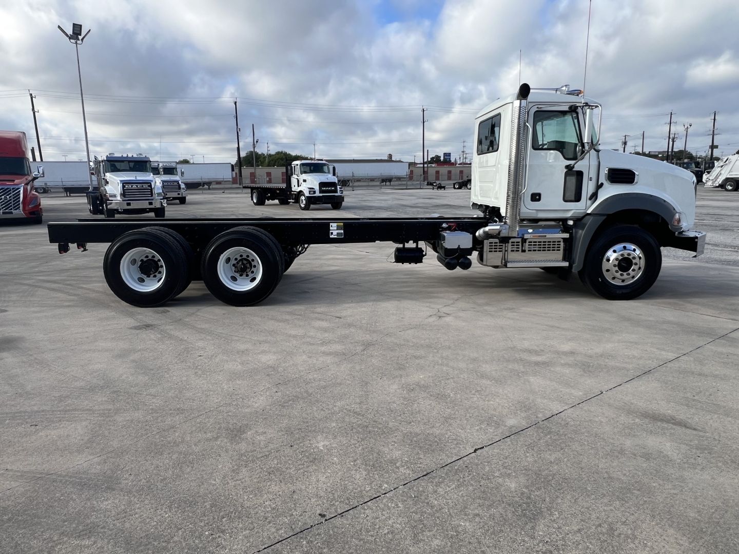 Mack GR64B cab and chassis truck side view showing flatbed with long wheelbase and dual rear axles; white cab, gray chassis, parking lot background.