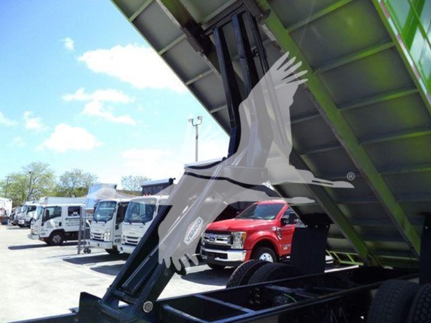Truck bed dump body hydraulic lifting mechanism visible with green support beam; multiple trucks in line in background.