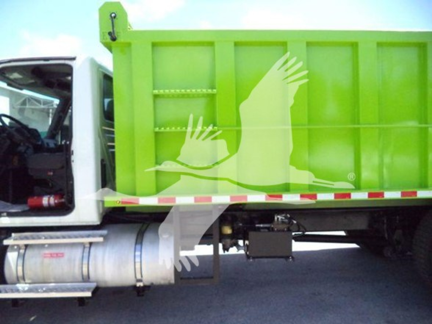 Truck side showing bright green dump bed with logo of flying birds, cab on left, visible fuel tank and undercarriage components.