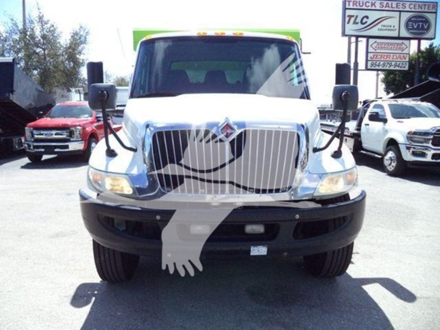 International dump truck front view with large chrome grille and emblem, headlights visible; text on dealership sign in background partially legible.