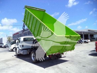 Dump truck with green dump bed raised, International MV truck bed in mid-tilt; site lot with other trucks in background.