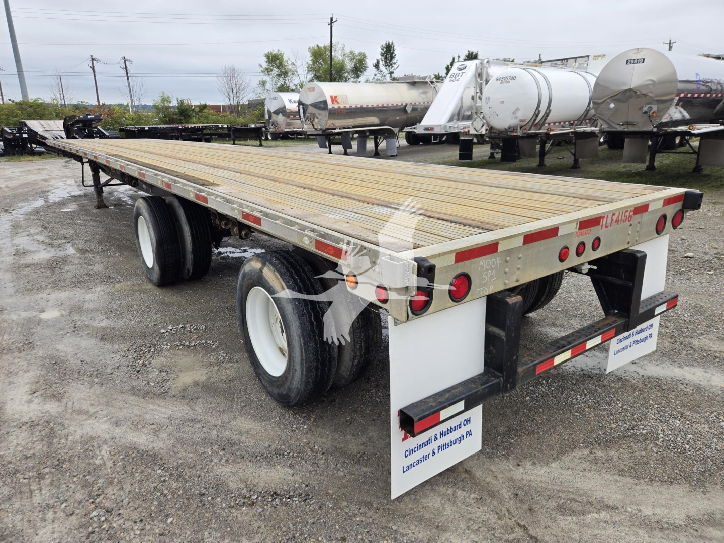 Flatbed trailer showing wooden deck, dual rear axles, and reflective safety stripes; utility equipment lot in background.