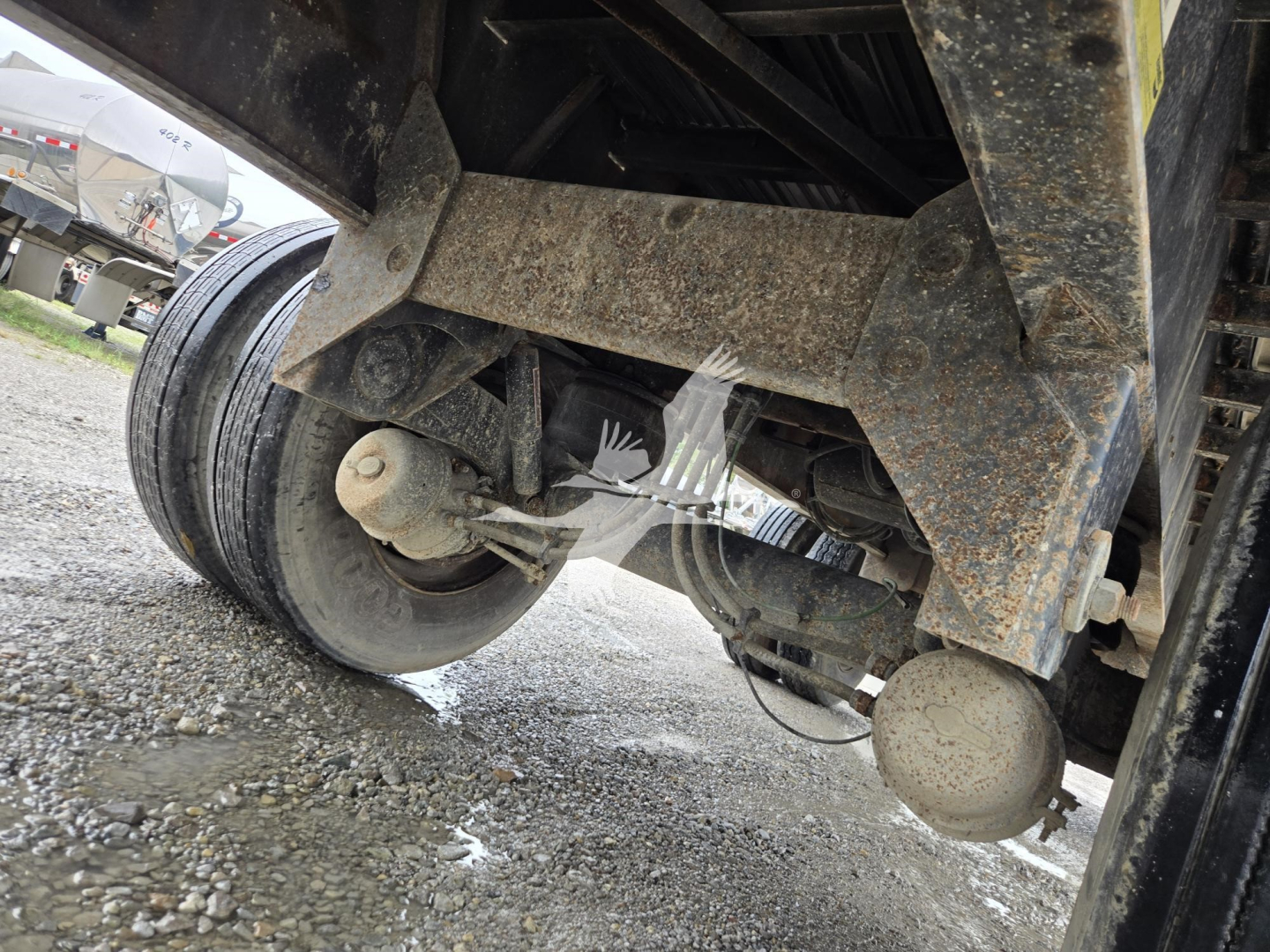Industrial trailer undercarriage showing axle assembly, leaf springs, and wheel hub with rust-like road grime; visible components include suspension members and wiring near the wheel.