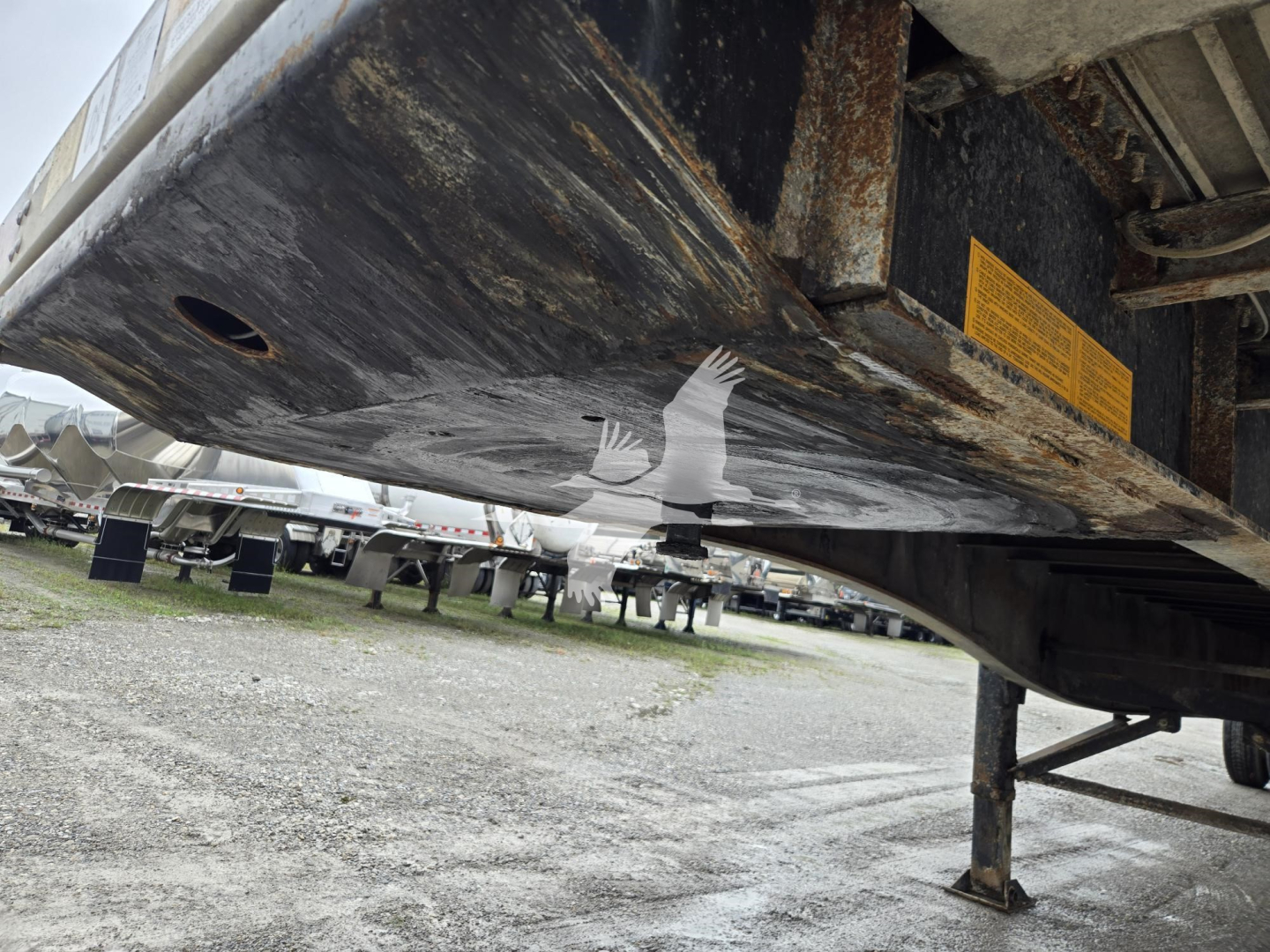 Trailer undercarriage with rust-stained frame showing yellow warning label and support jack in view.