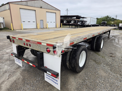 Flatbed trailer showing dual rear axles and wooden deck with reflective tape; rear bumper with lights and mudflaps; tires and wheel rims visible; industrial yard in background.