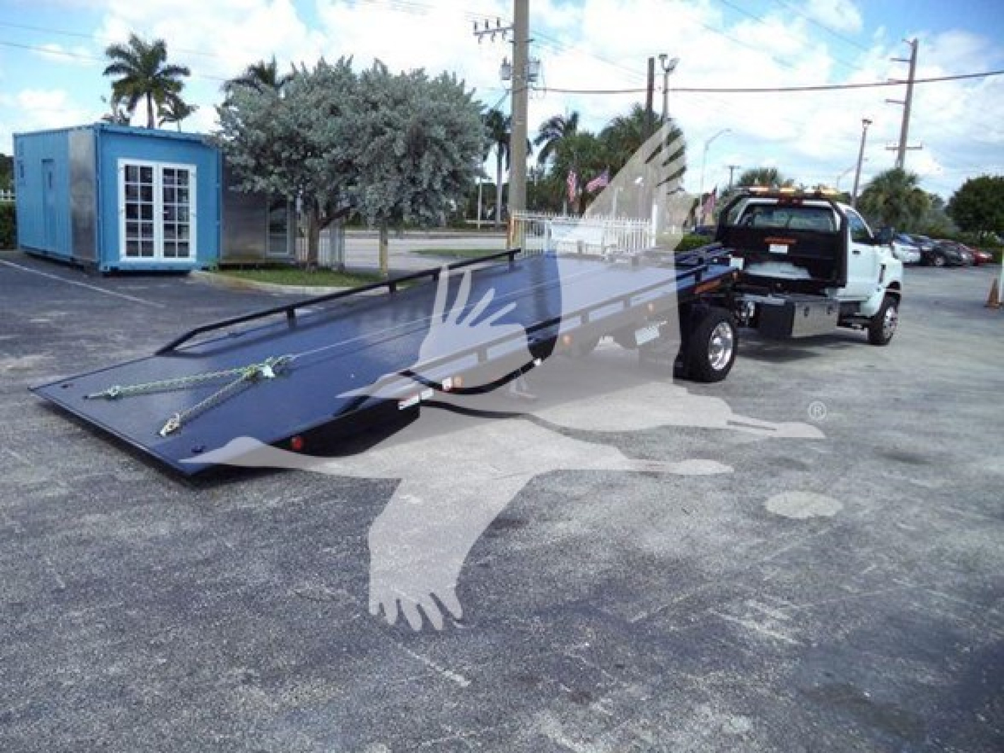 Flatbed tow truck with a blue cab and a long black ramp bed attached to a tow vehicle, driving on a paved lot with a blue portable building and palm trees in background.