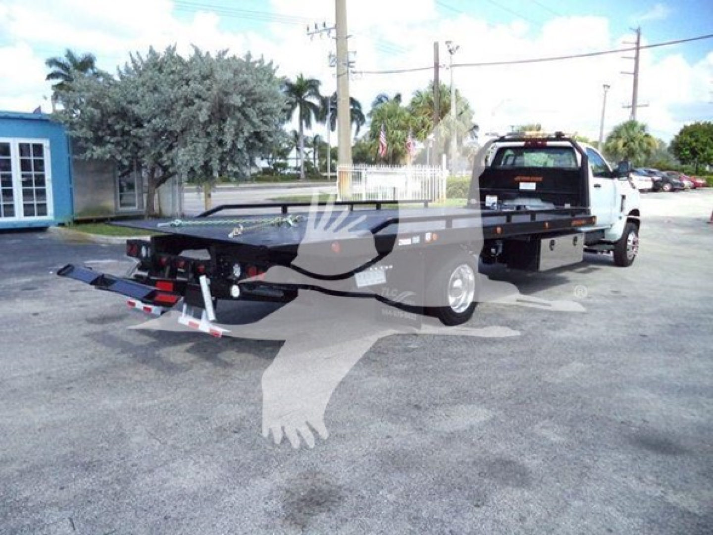 Flatbed tow truck in a dealership lot showing rear and long wheelbase, with hitch and ramps visible.
