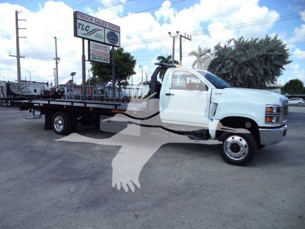 Flatbed tow truck in a dealership lot, white cab, long flatbed with equipment, signboards in background.