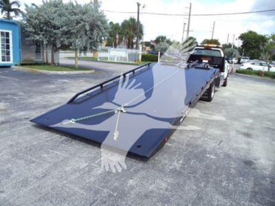 Truck rollback bed on a carrier with a long flatbed and ramp, image shows a black flatbed tow truck attachment with chains and wheel chocks, dealership lot in background.