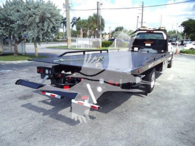 Flatbed tow truck with rear ramps and hitch, showing a corporate logo watermark on the image and a black flatbed under a pickup truck in an outdoor lot.