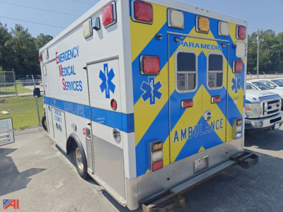 Rear view of a blue-and-yellow ambulance with 'AMBULANCE' and ems star of life symbols, red/blue emergency lights, and 'EMERGENCY MEDICAL SERVICES' text on side.