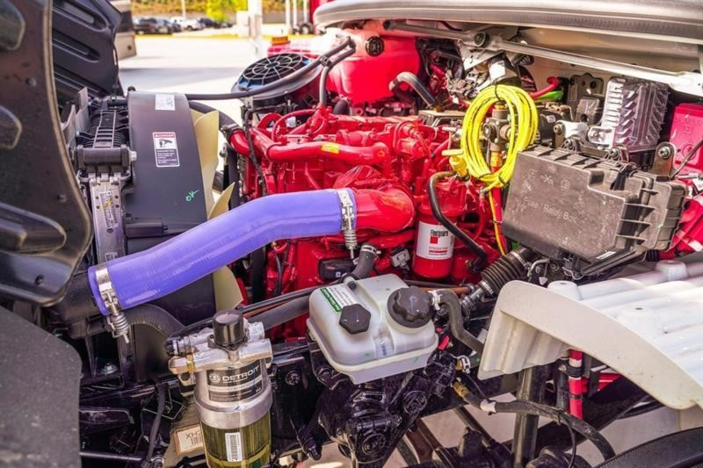 Freightliner M2 106 Plus engine bay showing red engine, blue coolant hose, yellow wiring, and various components in view.