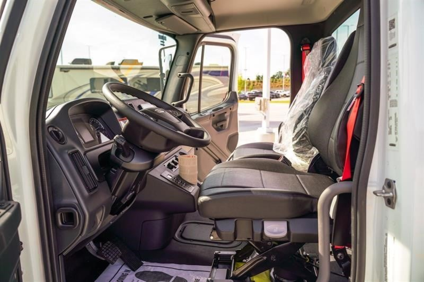 Interior view of Freightliner M2 106 Plus cab and chassis showing steering wheel, driver seat, and plastic-protected passenger seat.