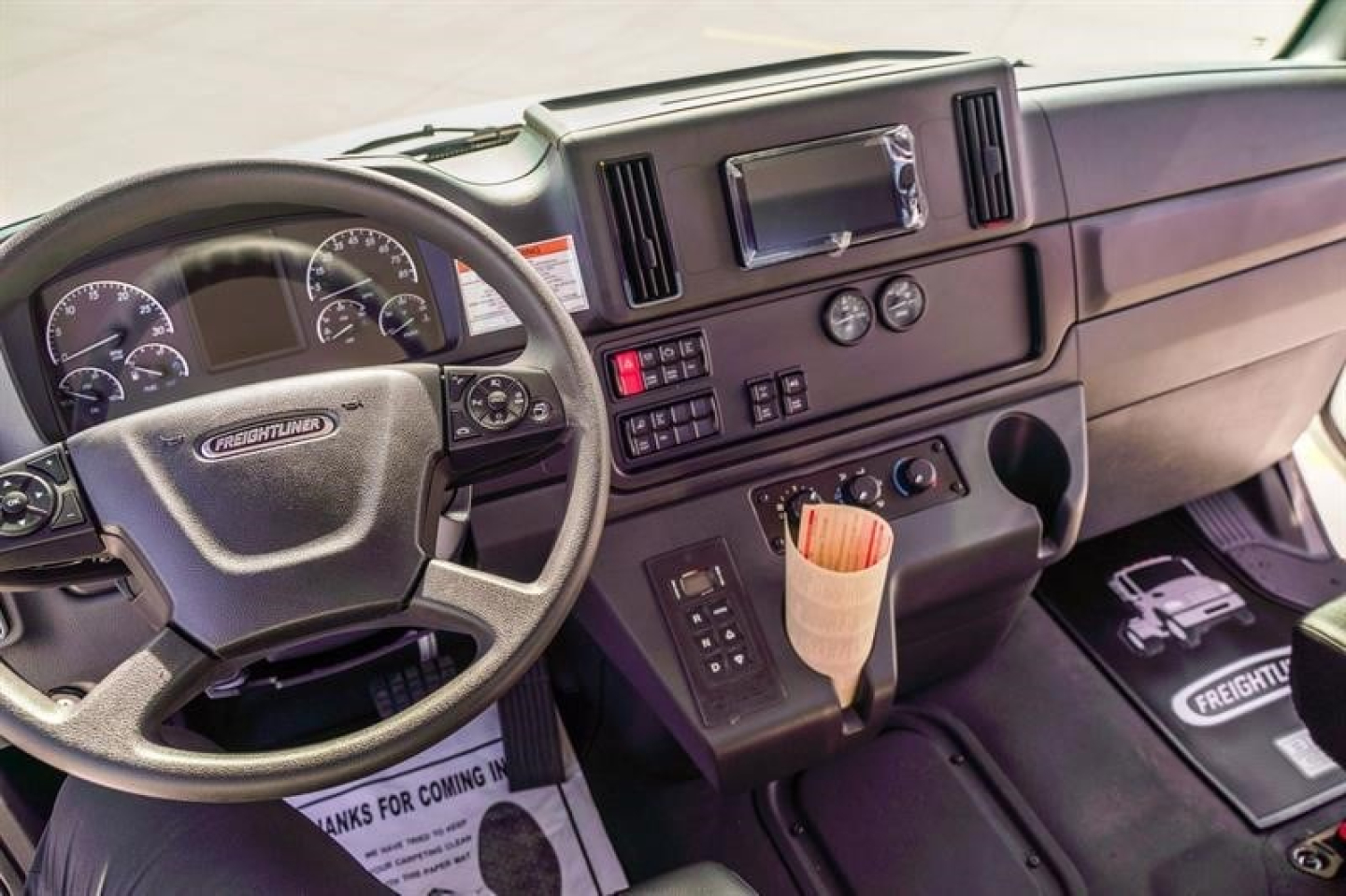 Cab and dashboard interior of a Freightliner truck showing steering wheel, instrument cluster, center console with controls, cup holder, and floor mat