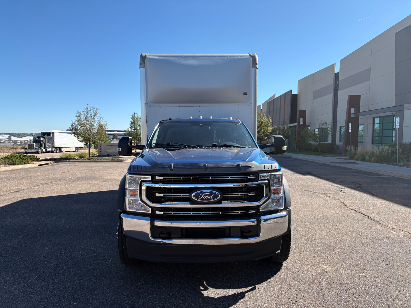 Blue Ford F-550 box truck front view with large cargo box, chrome grille, Ford emblem, in a parking lot beside a modern building.
