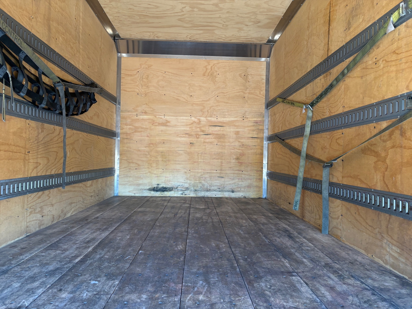 Truck cargo area interior showing wooden walls with metal E-track rails, straps hanging on left side, and a wooden floor.