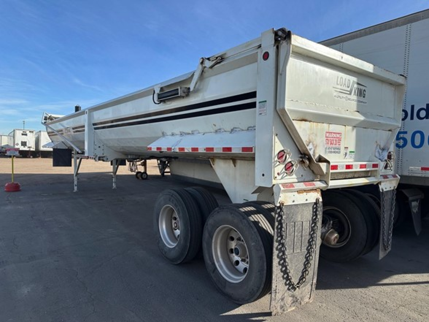 Load King rock end dump trailer side and rear showing hydraulic tailgate, white body with black stripe, dual rear axles, chain attachment, and warning decals.