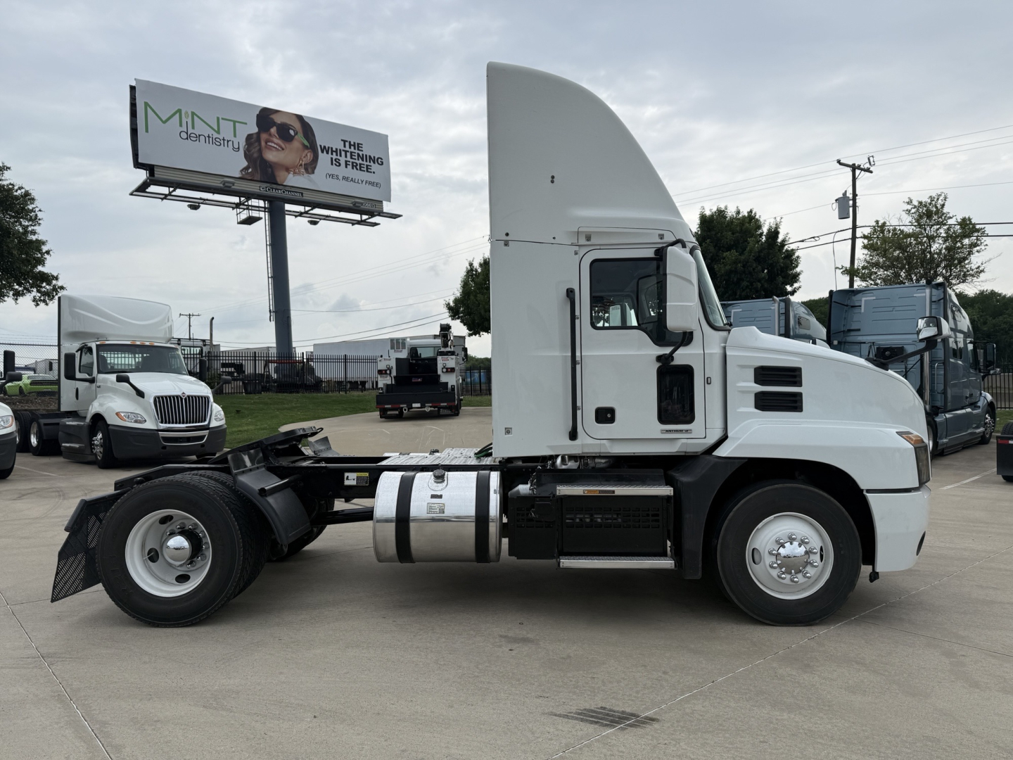 Truck. Side view of a white 2022 Mack AN42T conventional daycab tractor with gray fuel tank and exposed chassis; tall roof sleeper. Visible text on image signage in background.