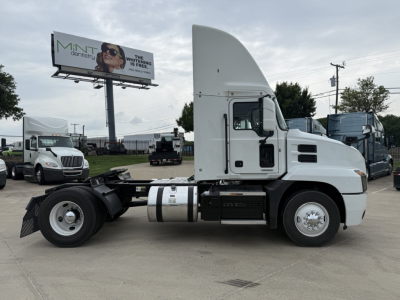 Truck. Side view of a white 2022 Mack AN42T conventional daycab tractor with gray fuel tank and exposed chassis; tall roof sleeper. Visible text on image signage in background.