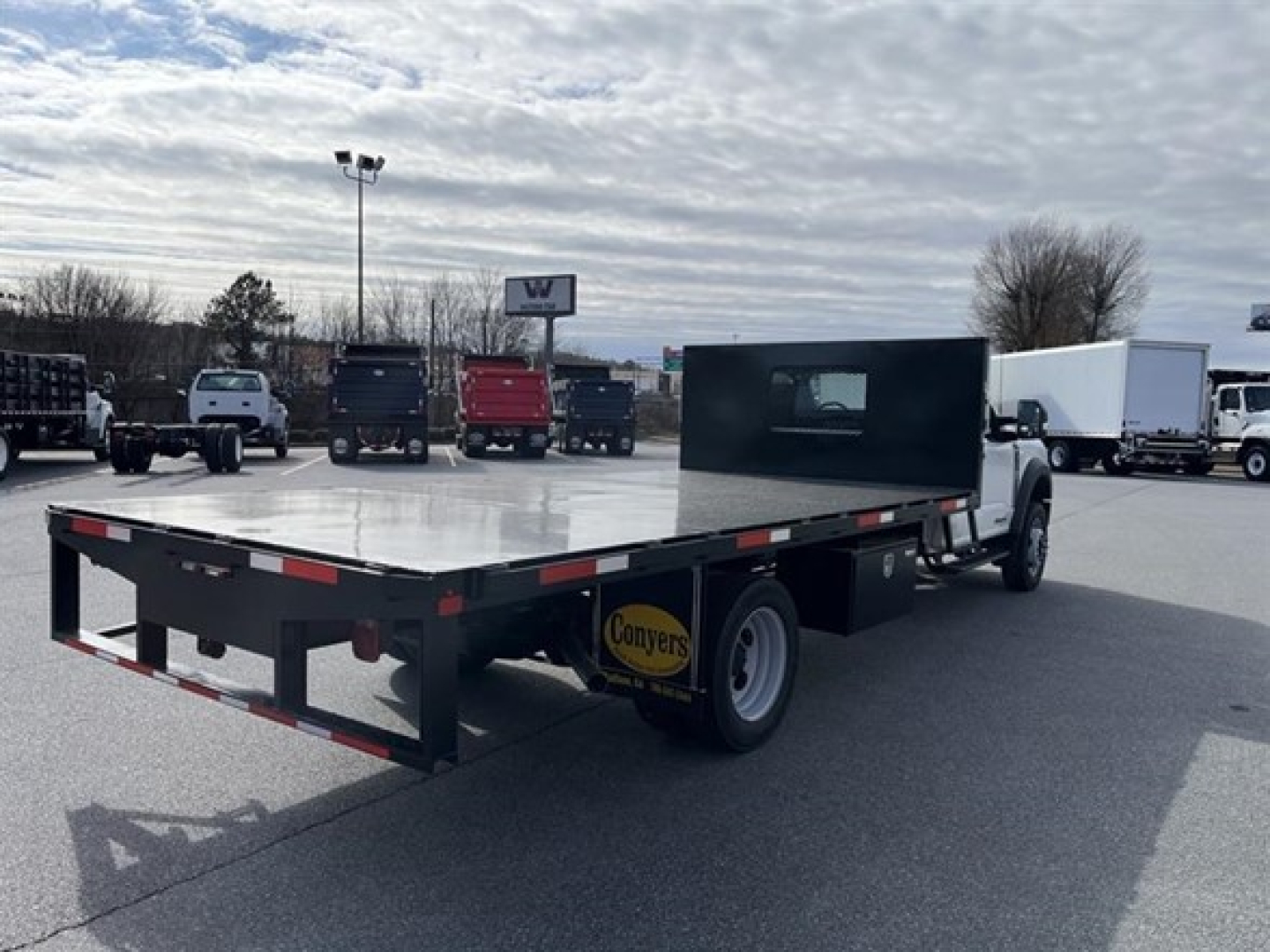 Flatbed tow truck chassis of a 2025 Ford F550 SD XL with a black flatbed, extended cab, and Conyers sticker on the front wheel area, seen in a dealership lot.