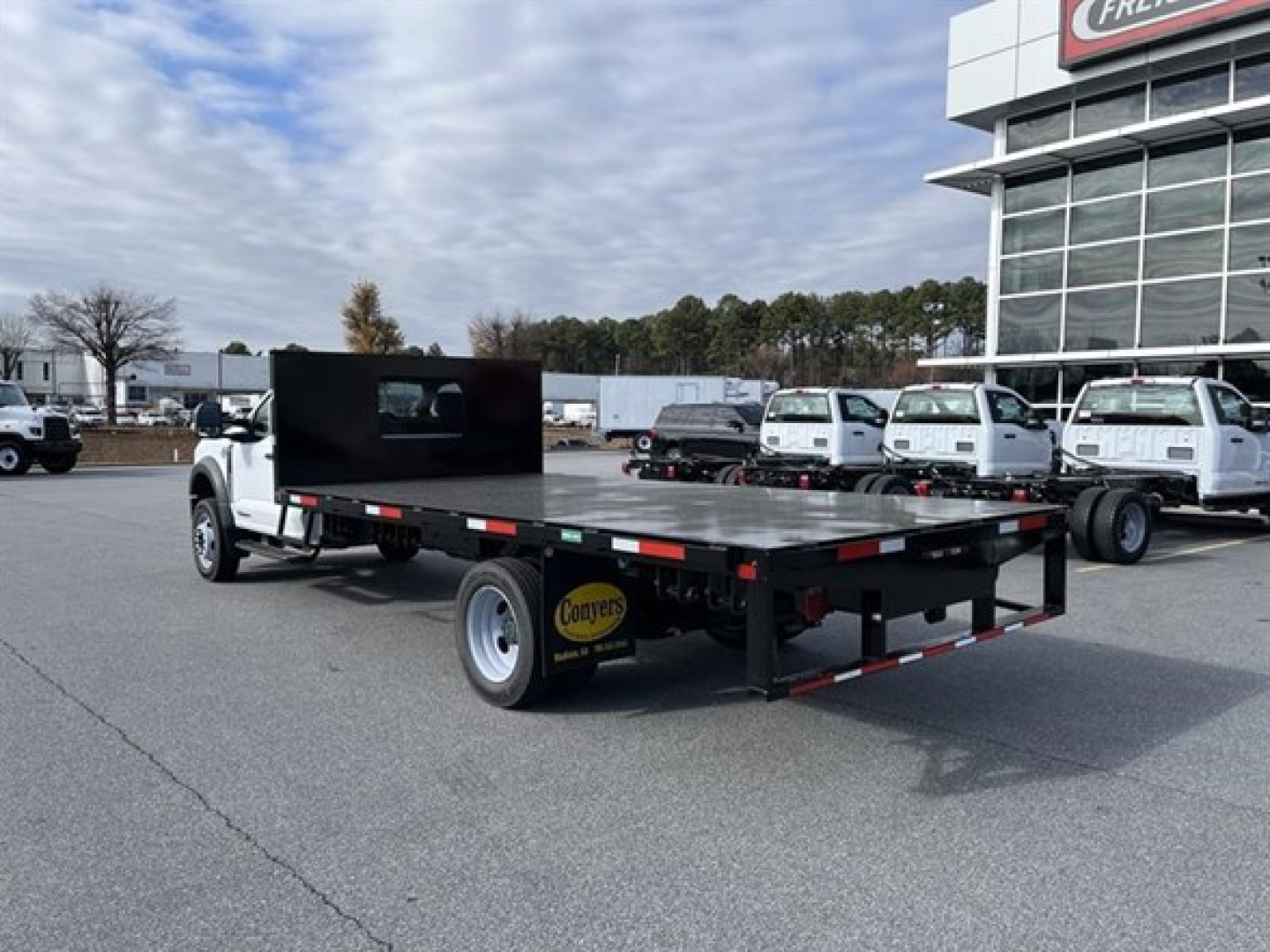 Flatbed tow truck on a dealership lot showing a long black flatbed with a divider screen and dual rear axle, white cab, and customers' fleet in background.