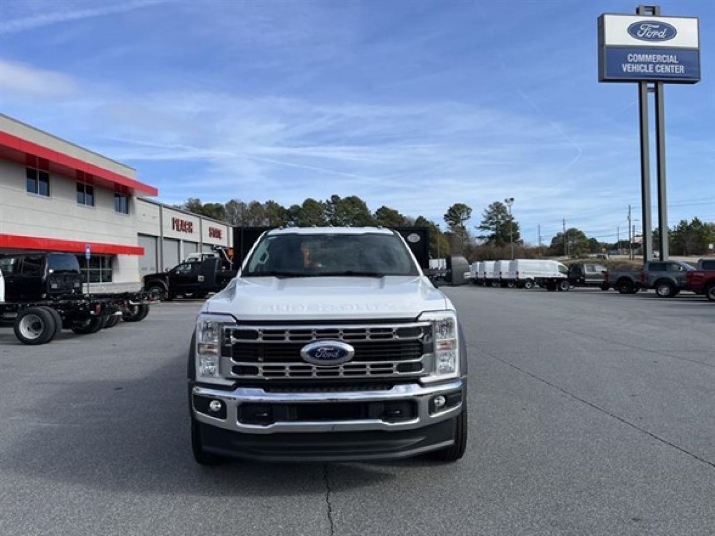 Front view of a white 2025 Ford F-550 SD XL flatbed truck parked in a dealership lot, Ford emblem on grille, daytime clear sky, other vehicles in background.