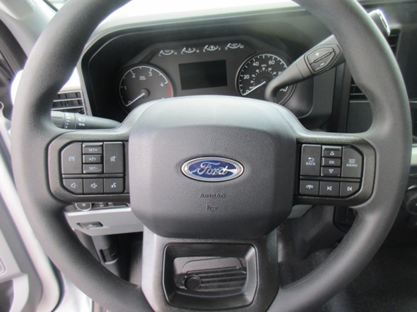 Ford steering wheel with control buttons in a truck cab, dash gauges visible behind, Ford emblem centered on steering wheel.