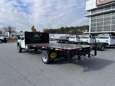 Flatbed tow truck on a dealership lot showing a long black flatbed with a divider screen and dual rear axle, white cab, and customers' fleet in background.