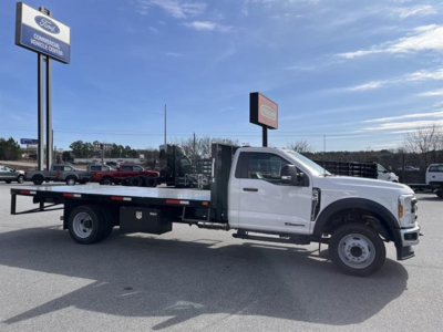 Ford F-550 flatbed truck in dealership lot, white cab with long flatbed, front wheels visible, dealership signs in background.
