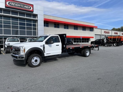 Flatbed tow truck in dealership lot showing white Ford F-550 SD XL with black flatbed and towing equipment, Freightliner dealership building in background.