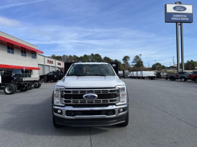 Front view of a white 2025 Ford F-550 SD XL flatbed truck parked in a dealership lot, Ford emblem on grille, daytime clear sky, other vehicles in background.