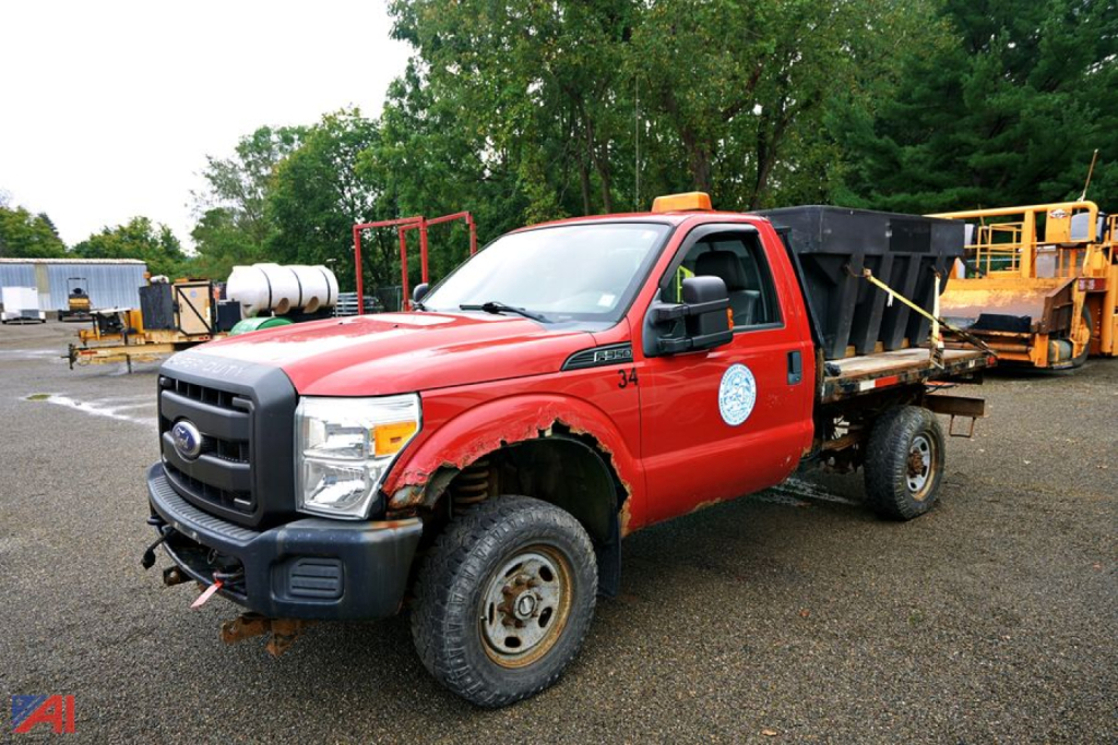Ford F-350 Super Duty flatbed truck with spreader attachment, red cab, black bumper, front view showing wheel well and utility equipment in background.