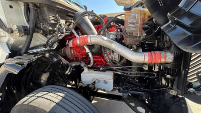 Truck engine bay with red engine block, silver intake pipe, and various hoses. Visible coolant reservoir, black chassis, and front wheel in view.