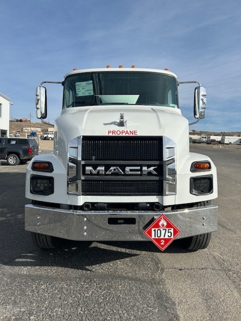 Front view of a white 2026 Mack MD7 truck showing large grille with MACK lettering, propane badge near windshield, orange clearance lights on roof, and a diamond hazard placard 1075 on bumper.