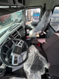 Truck cab interior of a new 2026 Mack MD7 showing steering wheel, dashboard, and plastic-covered seats with some paperwork and small boxes on the dash.