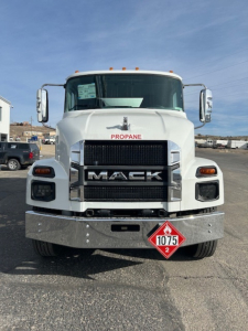 Front view of a white 2026 Mack MD7 truck showing large grille with MACK lettering, propane badge near windshield, orange clearance lights on roof, and a diamond hazard placard 1075 on bumper.
