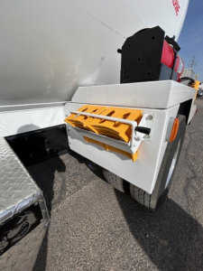 Wheel area of a Mack MD7 truck with a fuel or air tank and orange safety steps mounted on side, view of rear wheel and chassis.