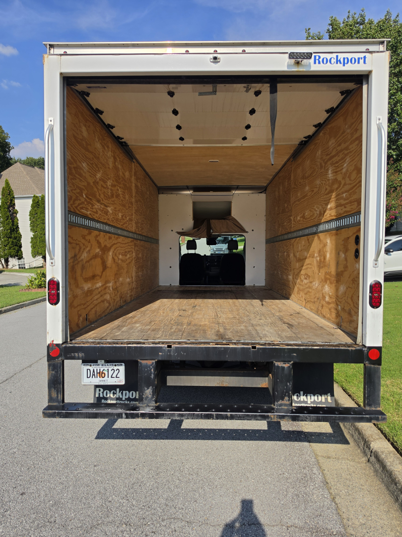 Truck cargo area showing empty box van interior with wooden walls and floor, rear doors open, Ford Transit 350 HD likely, Rockport branding on bumper.