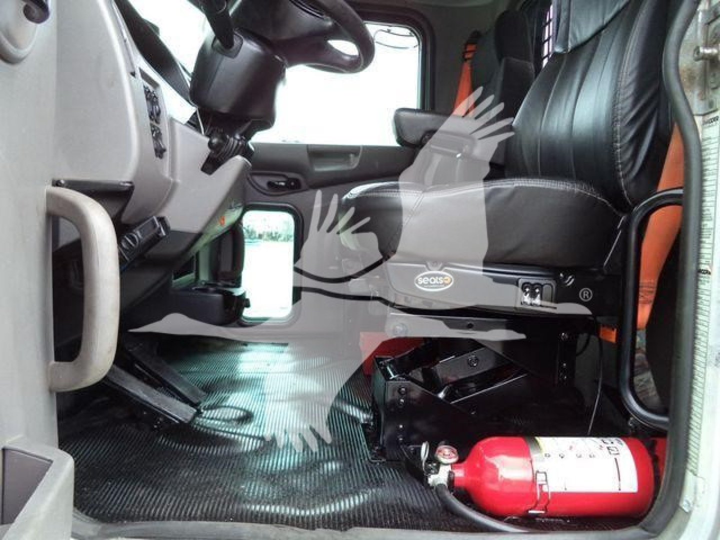 Truck cab interior with steering wheel, driver seat, and a red fire extinguisher on the floor. Dashboard area shows controls and a badge on the seat base.