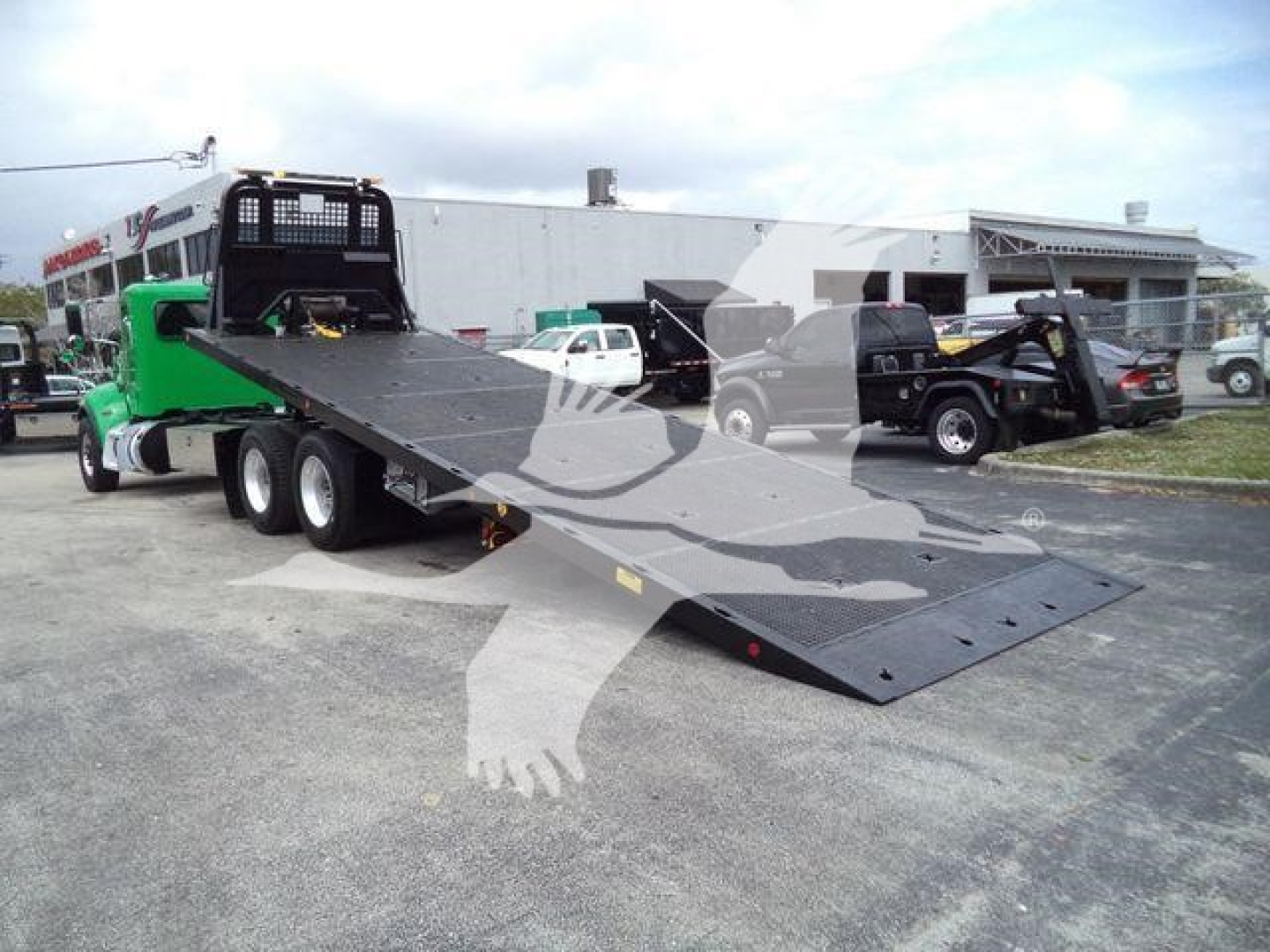 Truck with a green cab and a black rollback platform showing a long ramp in a parking lot.